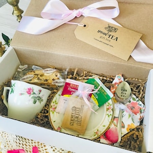 May include: A gift box with tea-related items. The box contains a teacup and saucer with floral designs, tea bags, a cookie, and a decorative spoon. The box is tied with a pink ribbon and has a tag that says "special TEA gift box."