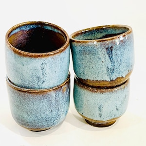 Puede incluir: Cuatro tazas de cerámica apiladas. Las tazas tienen un esmalte azul claro con bordes y bases marrones. El interior de las tazas es de un azul más oscuro. Las tazas están hechas a mano y tienen un aspecto rústico.
