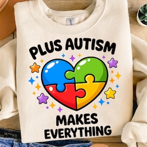 Op de afbeelding: Crèmekleurige sweatshirt met de tekst "PLUS AUTISM MAKES EVERYTHING" in zwart. Het ontwerp toont een hartvormige puzzel met blauwe, groene, rode en gele stukken, omringd door kleurrijke sterren. De sweatshirt ligt op een houten oppervlak.