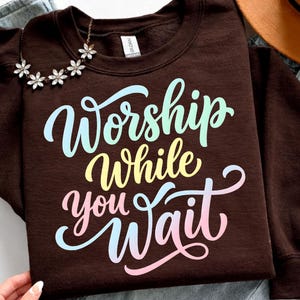 May include: A brown sweatshirt with the words "Worship While You Wait" in a colorful, script font. A floral necklace and a brown hat are also visible. The sweatshirt is made of a soft material.