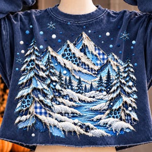 Puede incluir: Una sudadera azul marino de cuello redondo con una aplicación texturizada de un paisaje montañoso con pinos nevados, un río y copos de nieve. El diseño incorpora parches de tela con estampado de cuadros vichy y leopardo.