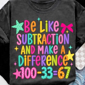 May include: A charcoal gray t-shirt featuring the colorful text "Be Like SUBTRACTION AND MAKE A DIFFERENCE 100-33=67". The design includes bright pink, yellow, orange, teal, and purple lettering, accented with star shapes and a pink bow graphic.