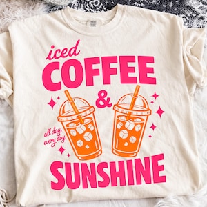 May include: A cream-coloured t-shirt with the words "iced COFFEE & SUNSHINE" in bright pink. Two orange iced coffee drinks with straws are featured below the text. The phrase "all day, every day" is printed below the coffee cups.