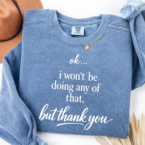 Puede incluir: Sudadera azul con cuello redondo con el texto "ok... I won't be doing any of that, but thank you" en escritura blanca. Un collar dorado con un colgante está sobre la sudadera. Un sombrero marrón es visible en el fondo.
