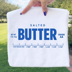 May include: A white t-shirt featuring a blue graphic print that mimics a butter wrapper. The text reads SALTED BUTTER, NET WT. 4 OZ. (113g), GRADE AA, and includes a measurement scale with markings for 1 TBSP, 1/4 CUP, 1/3 CUP, and 1/2 CUP.