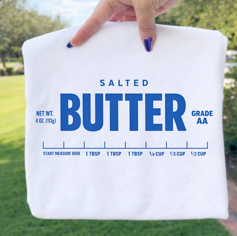 May include: A white t-shirt featuring a blue graphic print that mimics a butter wrapper. The text reads SALTED BUTTER, NET WT. 4 OZ. (113g), GRADE AA, and includes a measurement scale with markings for 1 TBSP, 1/4 CUP, 1/3 CUP, and 1/2 CUP.