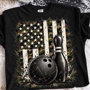 Może przedstawiać: Czarny t-shirt z krótkim rękawem z nadrukiem graficznym przedstawiającym kulę do kręgli i kręgiel umieszczone na tle stylizowanej flagi amerykańskiej. Wzór flagi zawiera kamuflaż z białymi gwiazdami i pionowymi pasami.
