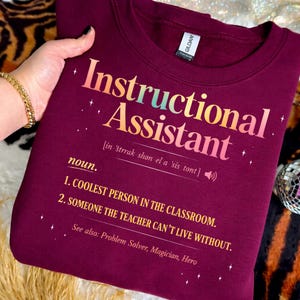 Puede incluir: Una sudadera granate con las palabras "Instructional Assistant" en una fuente de gradiente colorido. El texto incluye una definici&oacute;n, y la sudadera es sostenida por una persona.