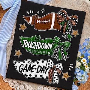 May include: A black rectangular sign featuring three horizontal banners with football-themed graphics. The top banner shows a brown football, the middle says TOUCHDOWN with yard lines, and the bottom says GAME DAY, all accented with bows and gold stars.