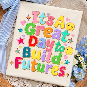 Könnte beinhalten: Eine weiße Stoffoberfläche mit dem bunten Text It's A Great Day to Build Futures in einer mehrfarbigen Blasen-Schriftart mit goldenen Glitzerumrandungen. Das Design enthält kleine rosa Herzen, gelbe Smileys und verschiedene mehrfarbige Sterne.