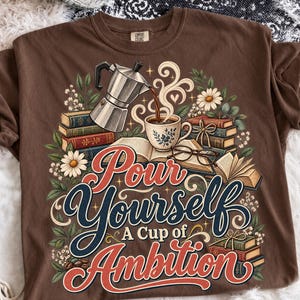 May include: A brown t-shirt featuring a graphic print of a silver moka pot pouring coffee into a cup, surrounded by stacks of books, eyeglasses, and white daisies. The text reads, Pour Yourself A Cup of Ambition, in stylized red and blue lettering.