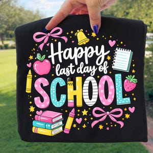 以下が含まれることがあります： 「Happy last day of SCHOOL」と書かれたカラフルなグラフィックが特徴の黒いTシャツ。ピンクのリボン、リンゴ、本、クレヨンなどのデザイン。Tシャツは手で持たれています。