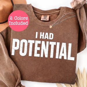 Pode incluir: Um moletom marrom com o texto "I HAD POTENTIAL" em letras brancas desgastadas. Um círculo rosa com o texto "2 cores incluídas" está no canto superior esquerdo. Um colar com pingente dourado é visível.