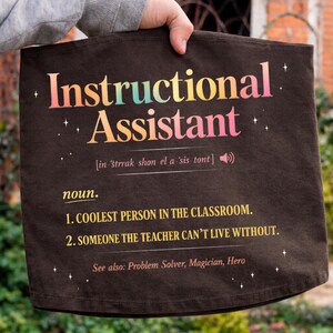 Puede incluir: Una bolsa de mano marr&oacute;n oscuro con las palabras "Instructional Assistant" en texto colorido. La bolsa incluye una definici&oacute;n, que enumera a la persona m&aacute;s genial del aula y a alguien sin quien el profesor no puede vivir.