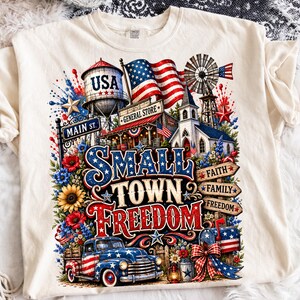 May include: A cream-coloured t-shirt featuring a graphic print of a small town scene with a water tower labelled USA, a general store, a church, a windmill, and a vintage pickup truck. The text reads Small Town Freedom, Faith, Family, Freedom.