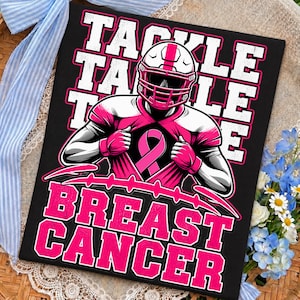 Op de afbeelding: Een zwart T-shirt met een afbeelding van een footballspeler in helm en shirt, die het shirt opentrekt om een roze lint voor borstkankerbewustzijn te tonen. De tekst TACKLE BREAST CANCER staat in grote witte en roze blokletters op het shirt.