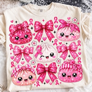 Może przedstawiać: Kremowy t-shirt z nadrukiem graficznym przedstawiającym kilka pierożków w stylu kawaii z uśmiechniętymi buźkami, ozdobiony jaskraworóżowymi brokatowymi kokardkami, małymi różowymi serduszkami, drobnymi kwiatkami i różowymi wstążkami świadomości.