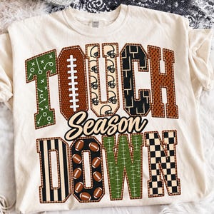 May include: A cream-colored t-shirt featuring the text TOUCHDOWN and the word Season in script. The letters are filled with various football-themed patterns including green play diagrams, brown football textures, helmets, and black checkers.