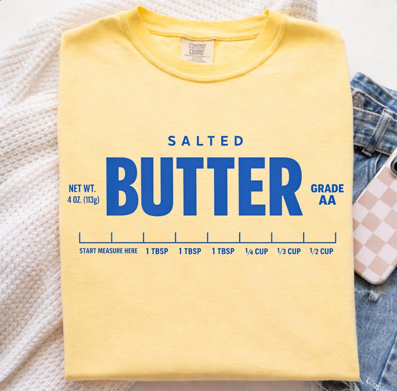 May include: A pale yellow t-shirt featuring blue text that reads SALTED BUTTER, NET WT. 4 OZ. (113g), and GRADE AA. Below the text is a blue measurement graphic with markings for 1 TBSP, 1/4 CUP, 1/3 CUP, and 1/2 CUP, labelled START MEASURE HERE.