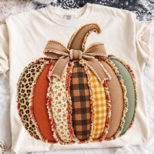 Puede incluir: Una camiseta de color crema con un gran gráfico central de una calabaza con múltiples patrones. La calabaza está compuesta por segmentos de tela verticales con estampado de leopardo, naranja sólido, floral, cuadros negros, vichy amarillo, beige y verde salvia.