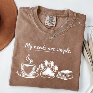 May include: Brown t-shirt with the text "My needs are simple." The graphic includes a coffee cup, paw print, and books. A silver necklace with a pendant is on the shirt. The shirt is folded on a white surface.