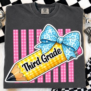 Könnte beinhalten: Ein anthrazitfarbenes T-Shirt mit einer Grafik eines gelben Bleistifts mit Karomuster, gekrönt von einer blauen Schleife mit Leopardenmuster. Der Bleistift zeigt den Text "Third Grade" in schwarzer Schrift vor vertikalen rosa Streifen.