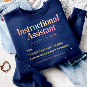 Puede incluir: Sudadera azul marino con la frase "Instructional Assistant" en letras coloridas. El texto incluye una definici&oacute;n: "La persona m&aacute;s genial del aula" y "Alguien sin quien el profesor no puede vivir."