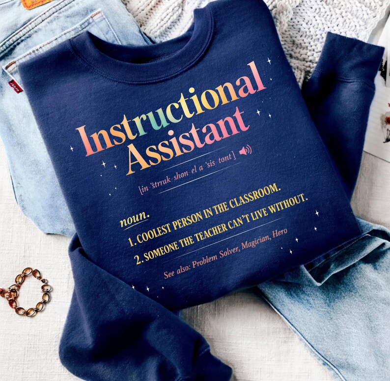 Puede incluir: Sudadera azul marino con la frase "Instructional Assistant" en letras coloridas. El texto incluye una definici&oacute;n: "La persona m&aacute;s genial del aula" y "Alguien sin quien el profesor no puede vivir."
