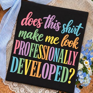 May include: A black t-shirt featuring the colorful text "does this shirt make me look PROFESSIONALLY DEVELOPED?" in a mix of pink, purple, light blue, green, yellow, and orange script and block lettering printed across the front of the fabric.