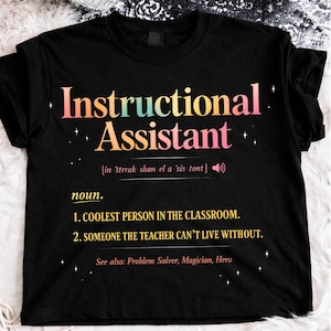 Puede incluir: Camiseta negra con la frase "Instructional Assistant" en una fuente de gradiente colorido. La camiseta incluye una definici&oacute;n, que indica que la persona es la "PERSONA M&Aacute;S GENIAL DEL AULA".