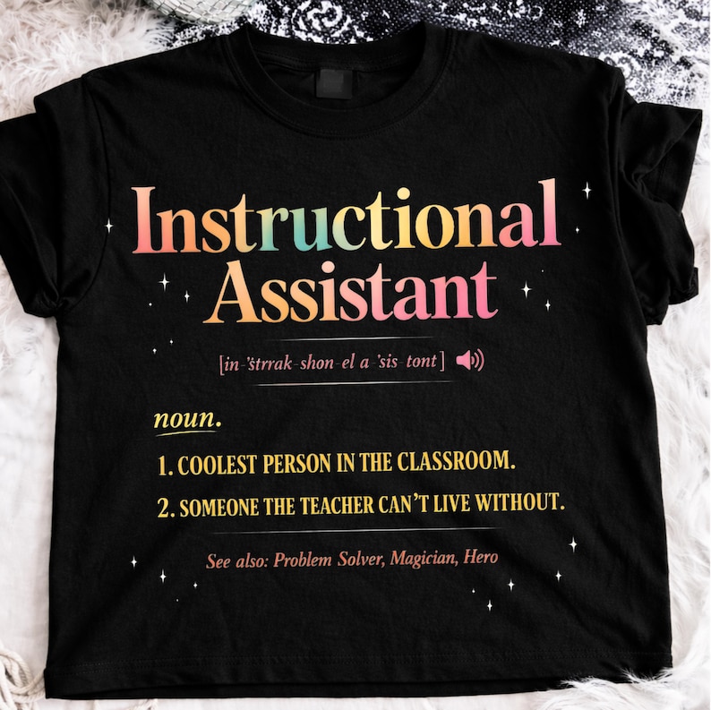 Puede incluir: Camiseta negra con la frase "Instructional Assistant" en una fuente de gradiente colorido. La camiseta incluye una definici&oacute;n, que indica que la persona es la "PERSONA M&Aacute;S GENIAL DEL AULA".
