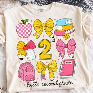 Può includere: Una t-shirt color crema con una stampa grafica raffigurante una mela a scacchi rosa, fiocchi gialli fatti di matite, una pila di libri, uno zaino rosa, un quaderno a spirale rosa con l'etichetta Note, il numero 2nd e il testo hello second grade.