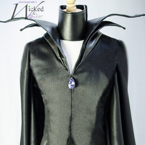 May include: Black costume with a high collar and a large, pointed neckline. The costume features a long, flowing cape and a decorative purple gemstone at the neckline. The costume is displayed on a mannequin.