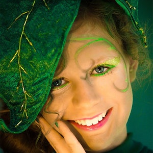May include: A person with green and gold makeup and a green leaf-shaped headpiece. The person is smiling, with green eyeliner and glitter eyeshadow. The headpiece has gold accents. The image has the text "Wicked & Wonder" and "© 2014 lane langmade photography".