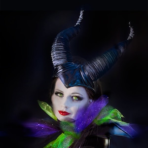 May include: A woman in a costume with a black horned headpiece, green eyes, and red lipstick. She wears a black outfit with a purple and green collar and holds a scepter. The image has a dark background.