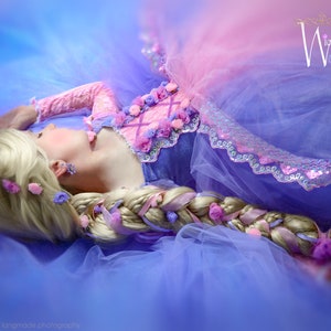 Pode incluir: Uma pessoa em um vestido rosa e roxo com longos cabelos loiros trançados adornados com fitas e flores. O vestido tem um corpete de renda e uma saia de tule. A imagem inclui o texto "Wicked & Wonder".