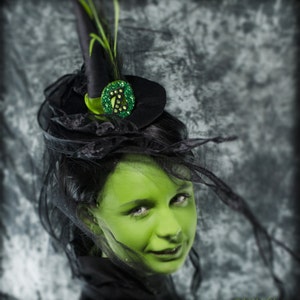 May include: A black witch hat with a green and black decorative accent, feathers, and a black veil. The person in the image has green skin and is wearing black clothing. The image has a Halloween theme.