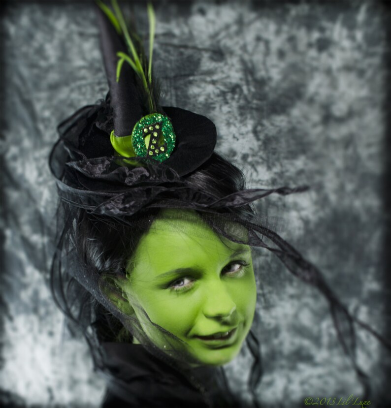 May include: A black witch hat with a green and black decorative accent, feathers, and a black veil. The person in the image has green skin and is wearing black clothing. The image has a Halloween theme.