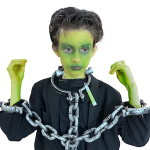 May include: A child dressed as Frankenstein for Halloween, with green face paint, a black jacket, and silver chain restraints around the neck and wrists. A glowing green stick hangs from the neck piece.