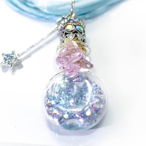 May include: A whimsical pendant necklace featuring a clear, round glass vial filled with iridescent glitter. The vial is adorned with a silver-toned crown and a pink crystal. A small silver wand with a star charm hangs beside the vial, all suspended from a light blue ribbon.