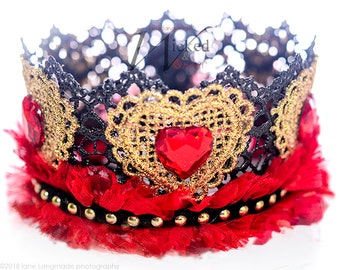 May include: A decorative crown with a black lace top, gold filigree, and a red heart-shaped jewel. The crown features red feathers and gold studs along the base, creating a regal and ornate design.