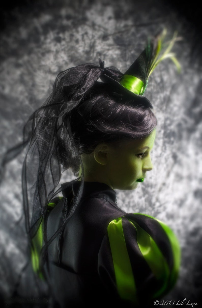 May include: A side profile of a person with green skin and green lipstick, wearing a black costume with green accents. The costume includes a black hat with a green ribbon and a black veil. The sleeves have green stripes.