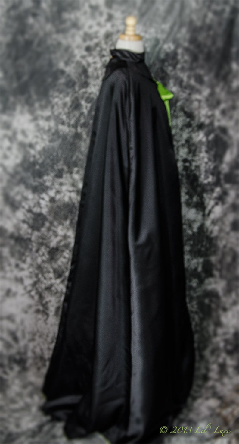 May include: A long, black cloak made of a shiny fabric, draped over a mannequin. The cloak has a high collar and a green accent near the neck. The background is a mottled gray and white. The copyright text "&copy; 2013 Lil' Luxe" is visible.