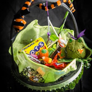 Może przedstawiać: Zielona, ​​tkana żaba siedzi w czarnym i zielonym koszu w kształcie kotła wypełnionym r&oacute;żnymi słodyczami na Halloween, w tym pudełkiem cukierk&oacute;w "Dots", pudełkiem cukierk&oacute;w "Jolly Rancher" i pudełkiem cukierk&oacute;w "Tootsie Roll".