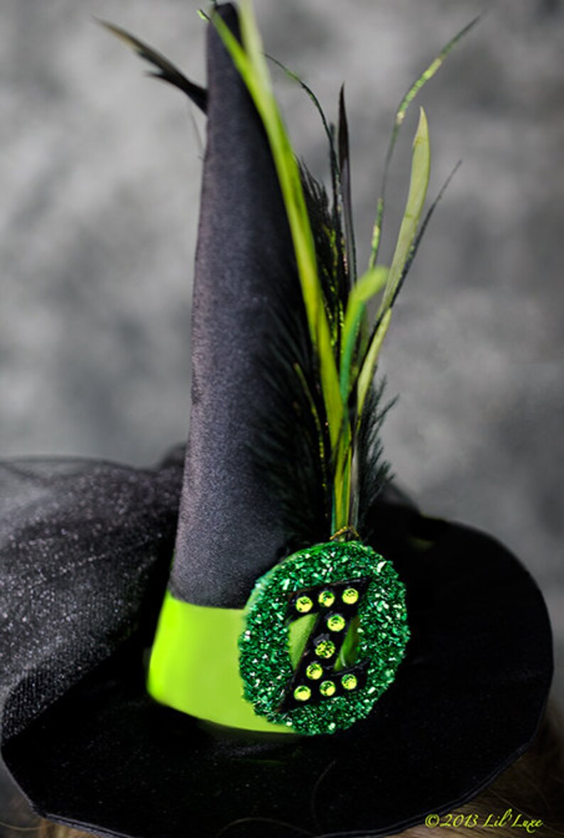 May include: A black witch hat with a bright green band and a decorative green glitter emblem. The emblem features the letter "Z" and is adorned with green gems. Black and green feathers are attached to the hat.