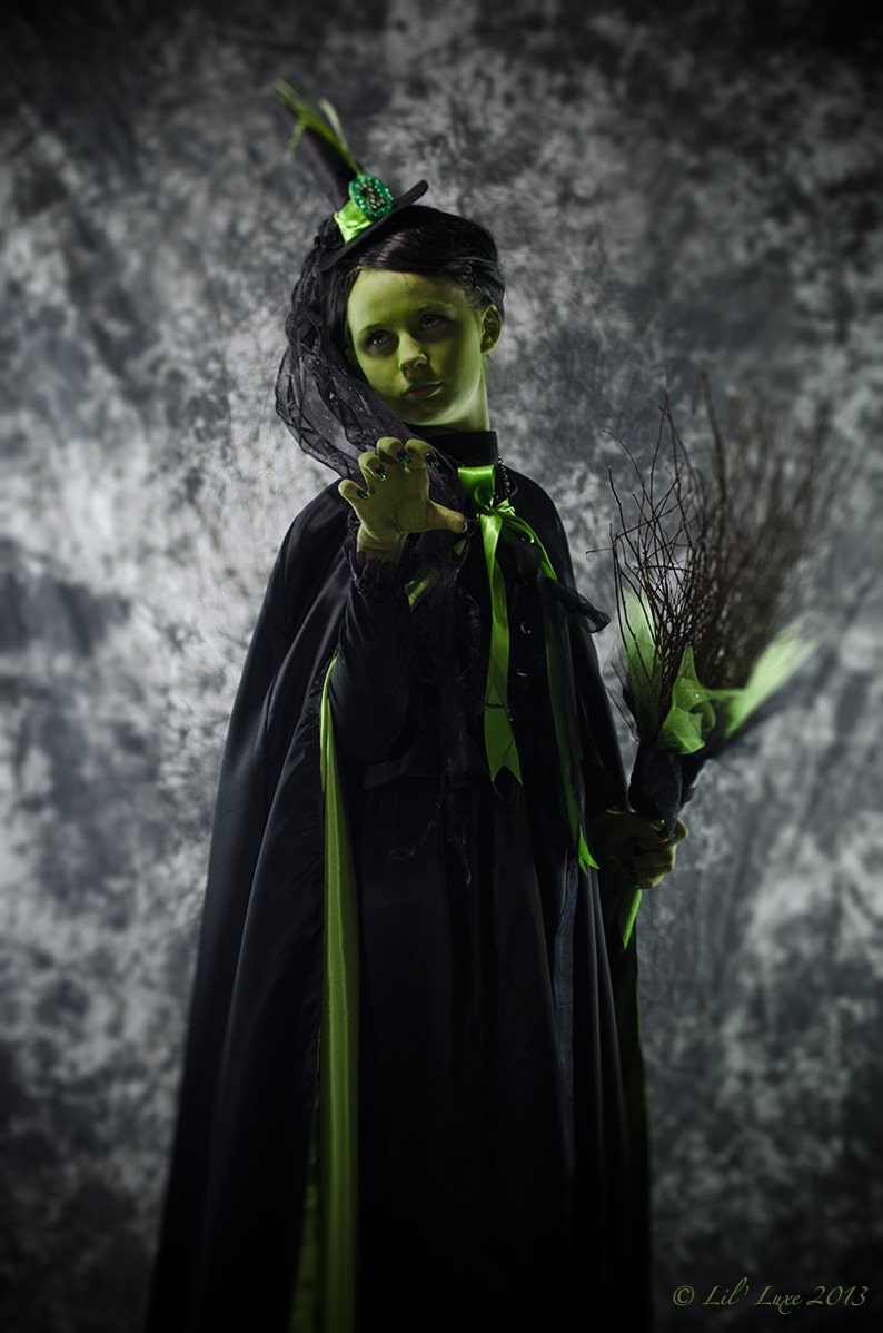 May include: A person in a witch costume with green skin, wearing a black dress with green accents and a black cape. They hold a broom and wear a black hat with a green jewel. The background is a mottled gray.