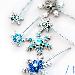 May include: Close-up of several snowflake hair pins. The pins have silver metal snowflake designs with various blue and clear crystal accents. The pins are arranged on a white surface, with some in focus and others blurred.