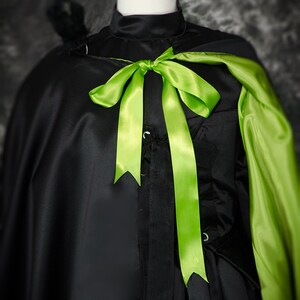 May include: Black satin cape with a high collar and a bright green satin bow. The cape has a green lining and is displayed on a mannequin. A black feather detail is visible on the shoulder.