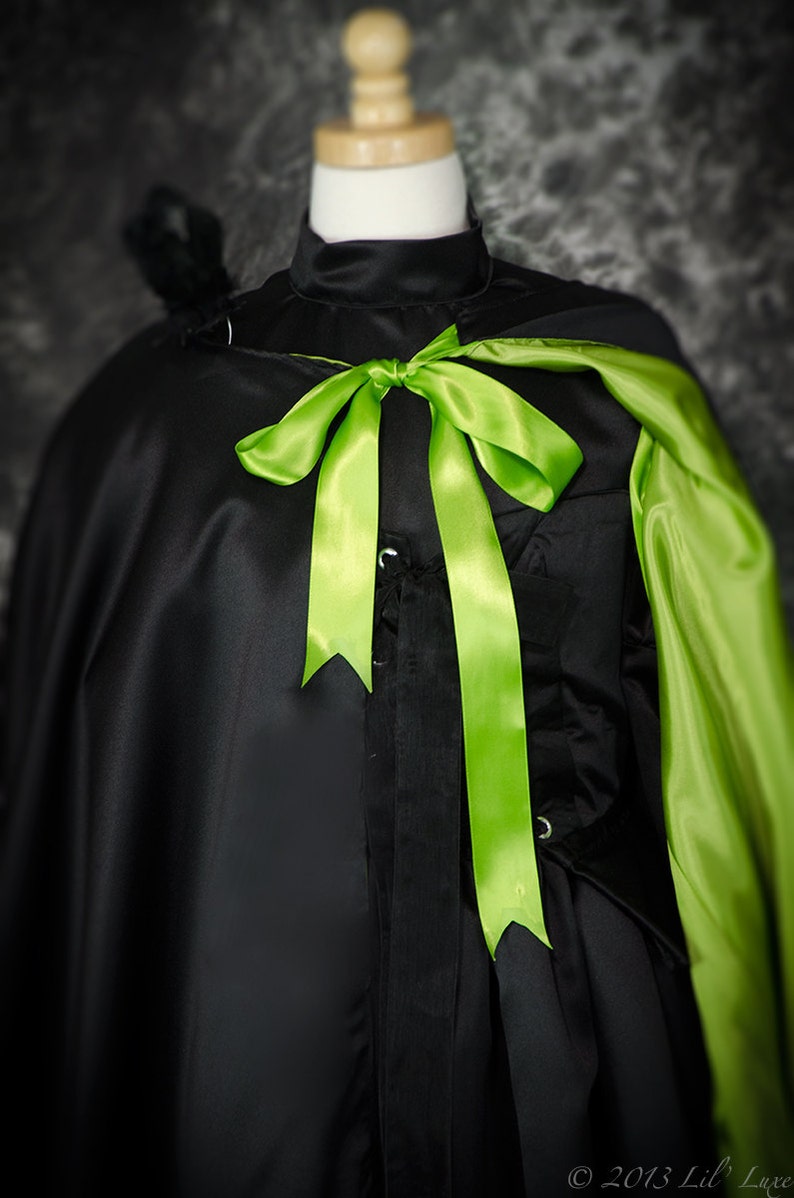 May include: Black satin cape with a high collar and a bright green satin bow. The cape has a green lining and is displayed on a mannequin. A black feather detail is visible on the shoulder.