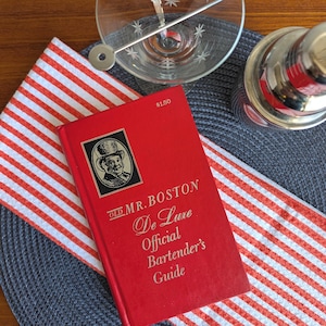 May include: A red "Old Mr. Boston De Luxe Official Bartender's Guide" book with gold lettering and a black and white illustration. A cocktail shaker, a martini glass with starburst etchings, and a bar spoon are also visible.
